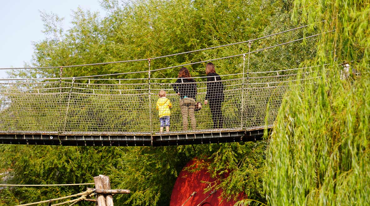 two adults and a child in a yellow coat on suspension bridge above the green park amongst the trees